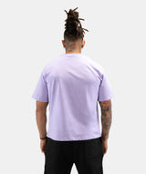 A man viewed from behind wearing a light lavender crewneck t-shirt and black pants, standing against a solid light gray background.