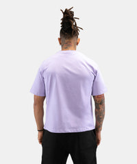 A man viewed from behind wearing a light lavender crewneck t-shirt and black pants, standing against a solid light gray background.