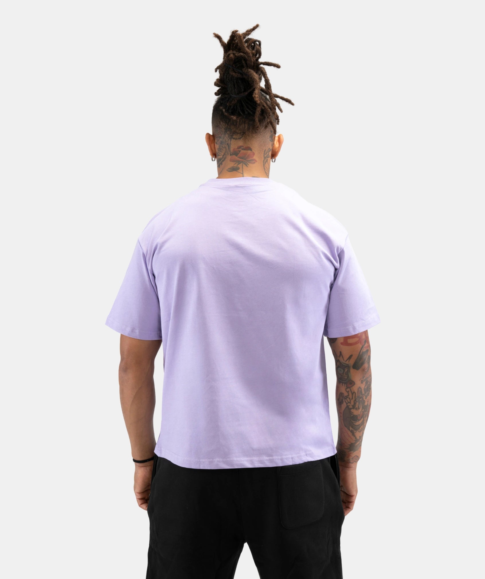 A man viewed from behind wearing a light lavender crewneck t-shirt and black pants, standing against a solid light gray background.