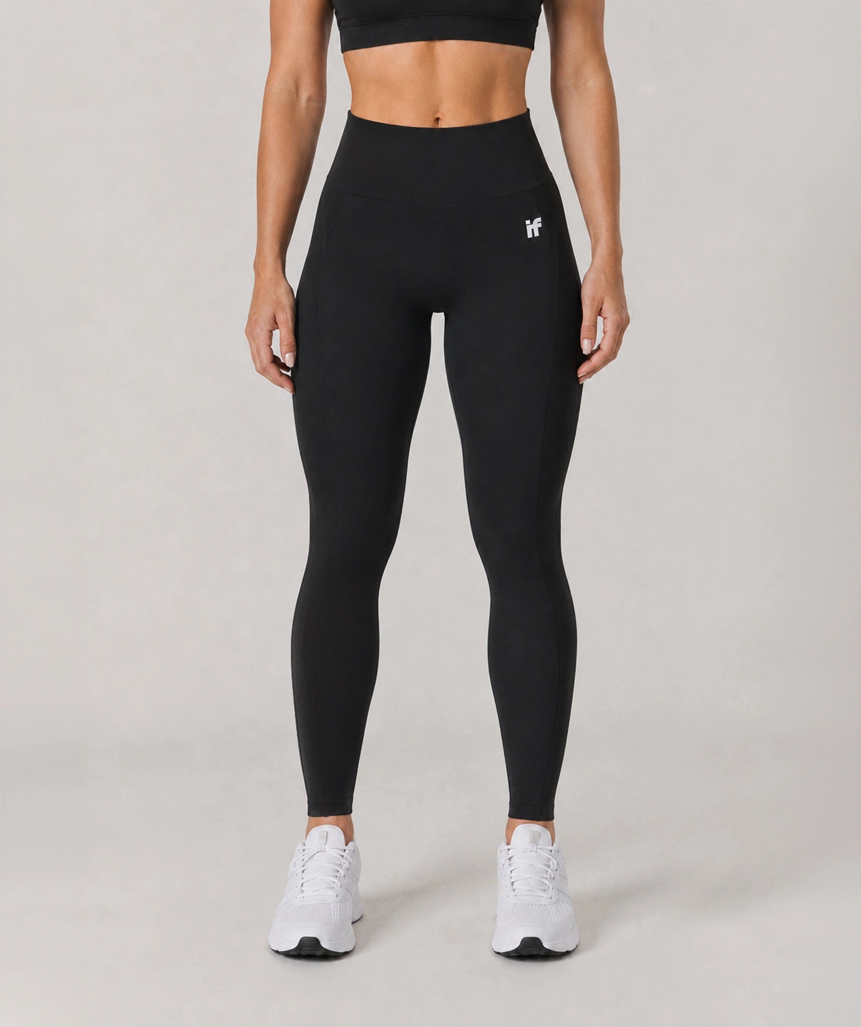 A model from the waist down wearing black high-waisted leggings with a small white logo on the upper left hip, paired with white sneakers, against a neutral light gray studio background.