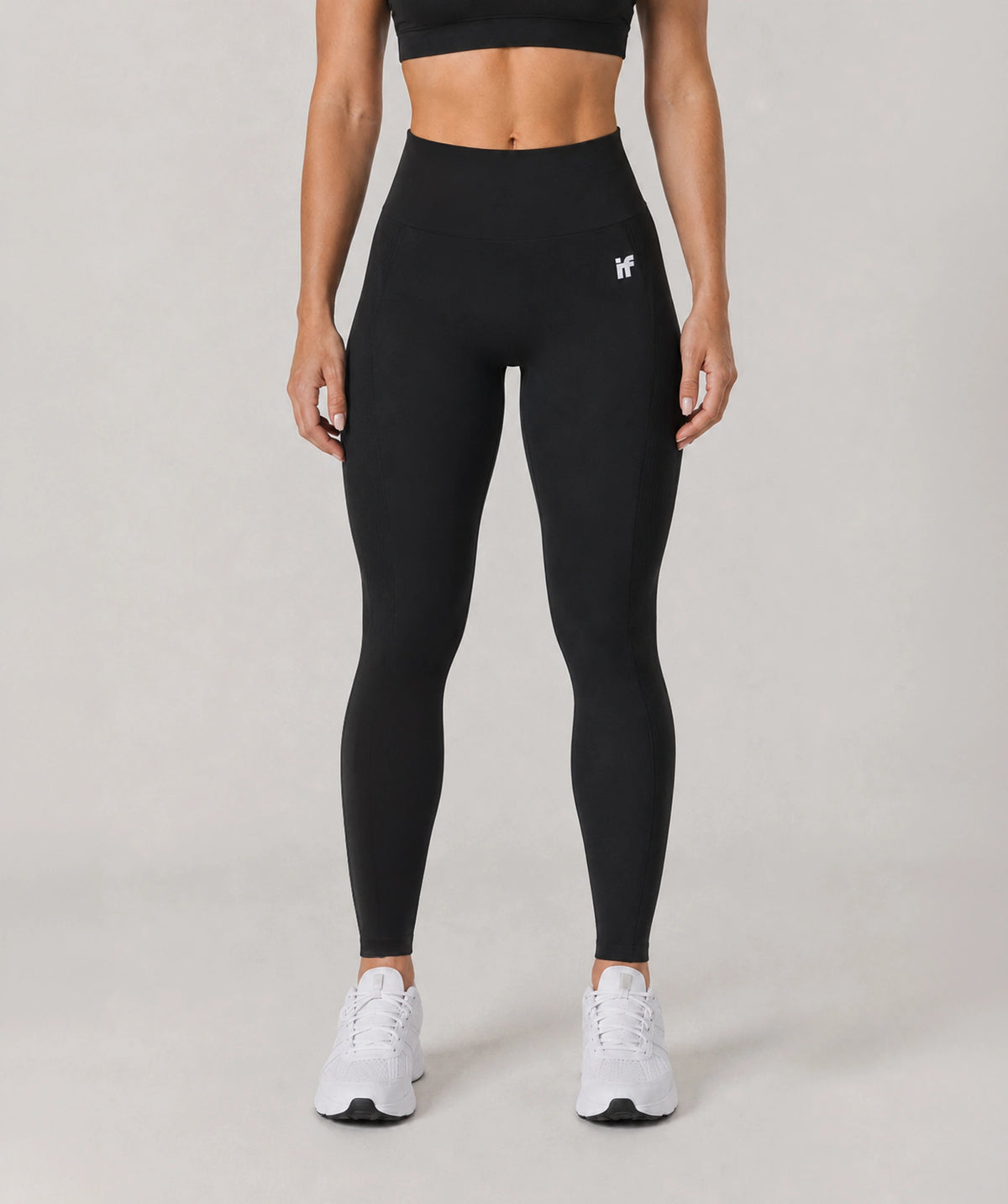 A model from the waist down wearing black high-waisted leggings with a small white logo on the upper left hip, paired with white sneakers, against a neutral light gray studio background.
