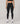 A model from the waist down wearing black high-waisted leggings with a small white logo on the upper left hip, paired with white sneakers, against a neutral light gray studio background.