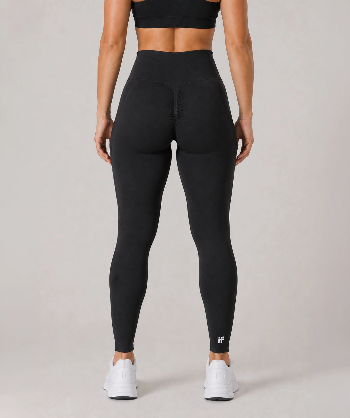 Rear view of a model wearing black high-waisted leggings, a black sports bra, and white sneakers against a light gray studio background.