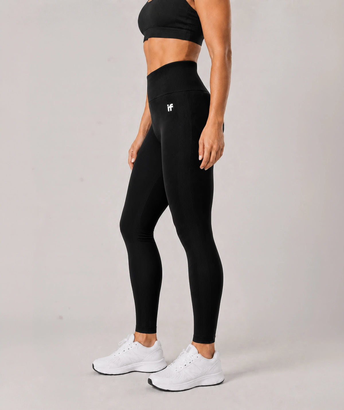 A woman modeling black high-waisted leggings with a small white logo on the left hip, paired with a black sports bra and white sneakers, against a light gray studio background.