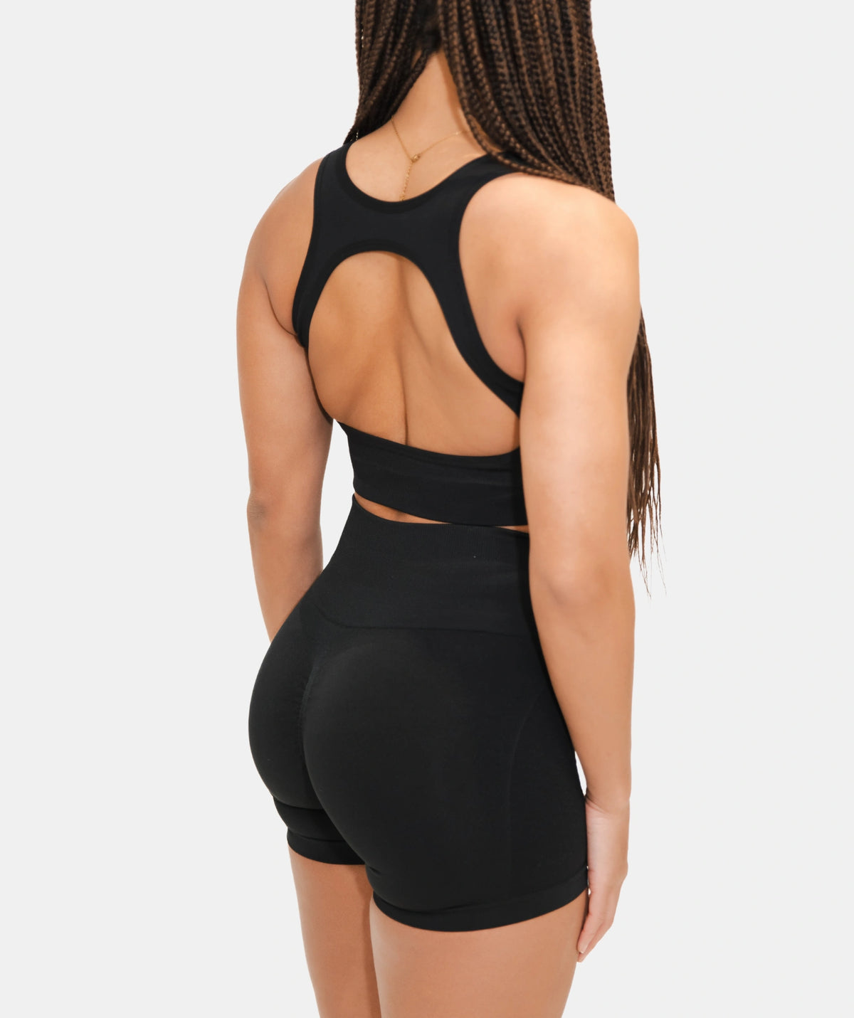 Rear view of a woman wearing a black athletic two-piece set with a cutout back sports bra and high-waisted shorts, against a light gray studio background.