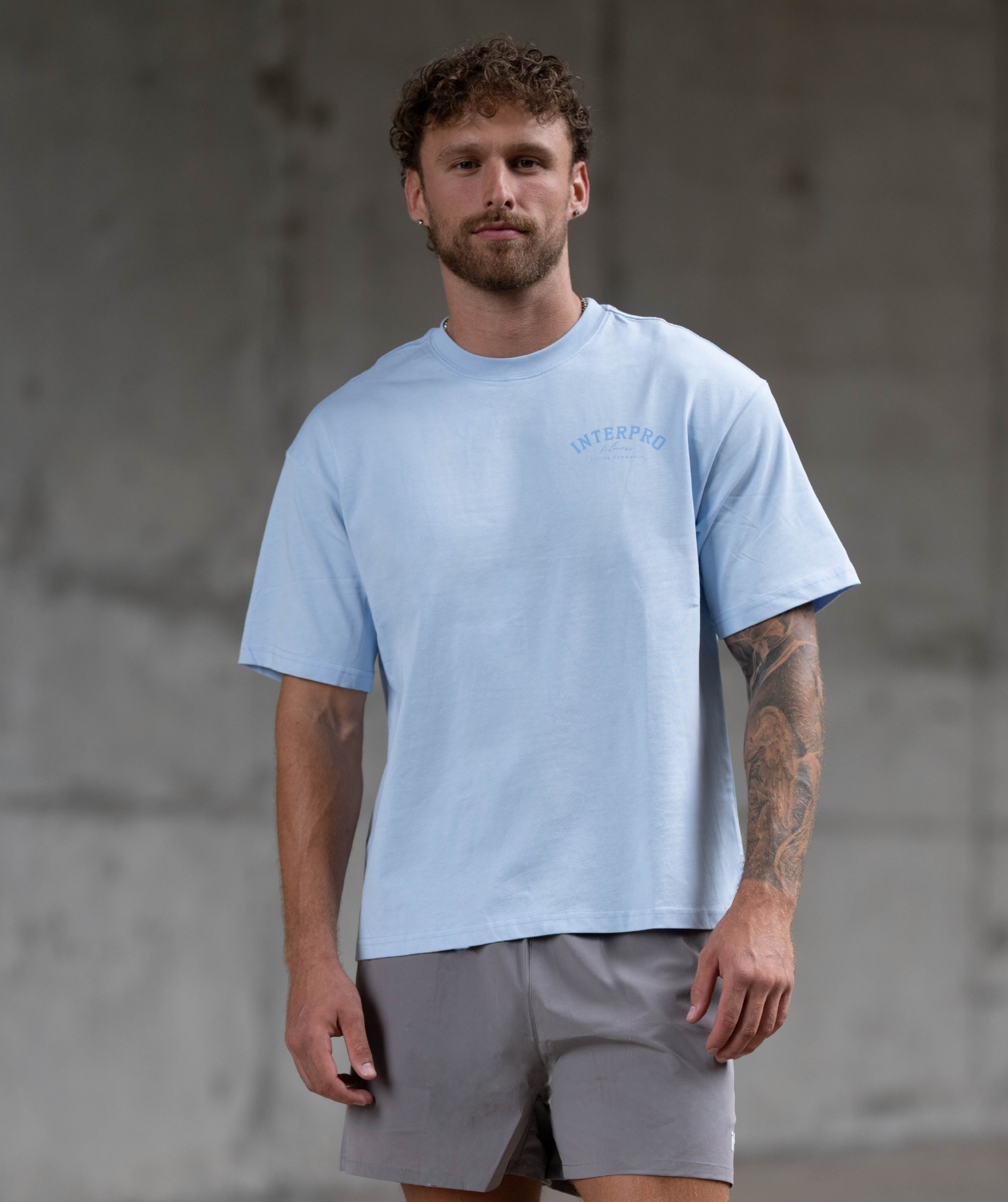 Man wearing a light blue Interpro Fitness t-shirt and grey shorts against a concrete wall. Front view