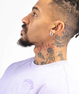 A close-up side profile of a man with neck and ear tattoos wearing a light lavender crewneck t-shirt with a purple graphic print on the chest.