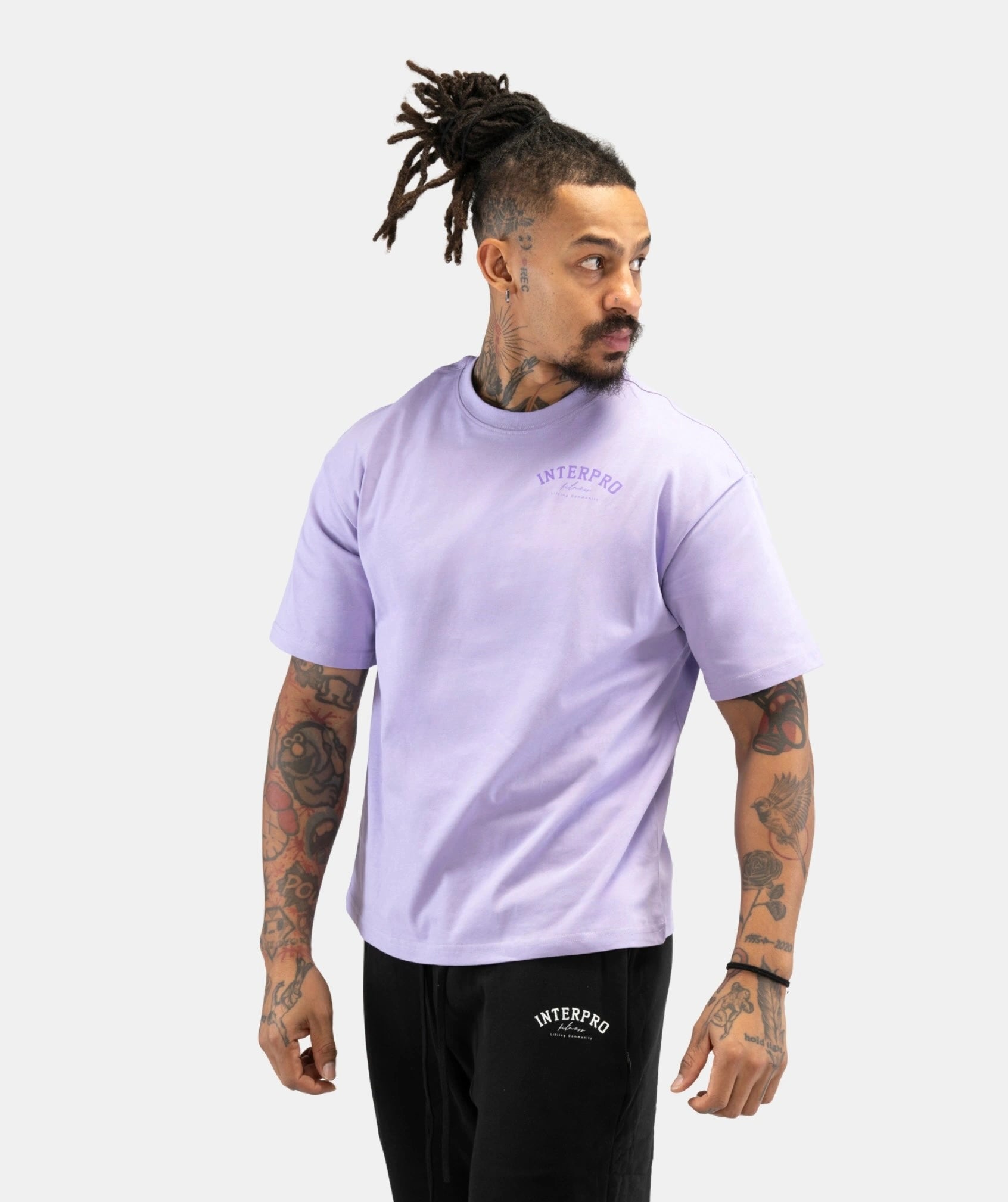 A man with dreadlocks modeling a light purple short-sleeve t-shirt with a small purple 'INTERPRO' logo on the left chest, paired with black sweatpants featuring a white logo on the thigh, against a light gray background.
