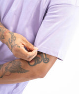 Close-up of a person wearing a light lavender short-sleeve t-shirt, pulling the sleeve up with one hand. The person has detailed black and gray tattoos on both arms, and the background is solid white.