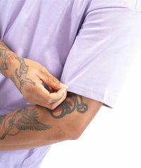 Close-up of a person wearing a light lavender short-sleeve t-shirt, pulling the sleeve up with one hand. The person has detailed black and gray tattoos on both arms, and the background is solid white.