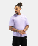 A man with dreadlocks and tattoos wearing a light purple t-shirt and black sweatpants, standing against a light gray background.