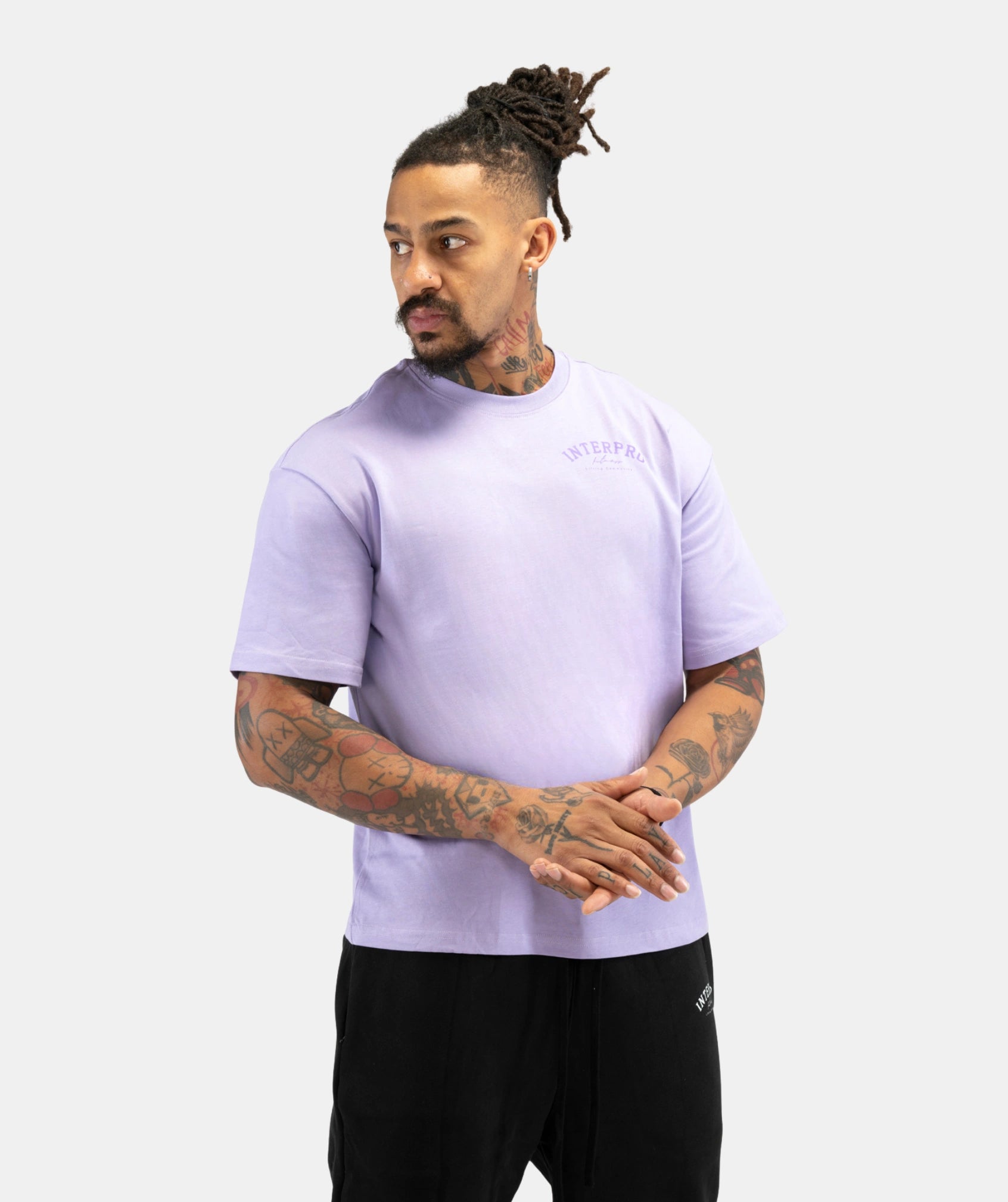 A man with dreadlocks and tattoos wearing a light purple t-shirt and black sweatpants, standing against a light gray background.