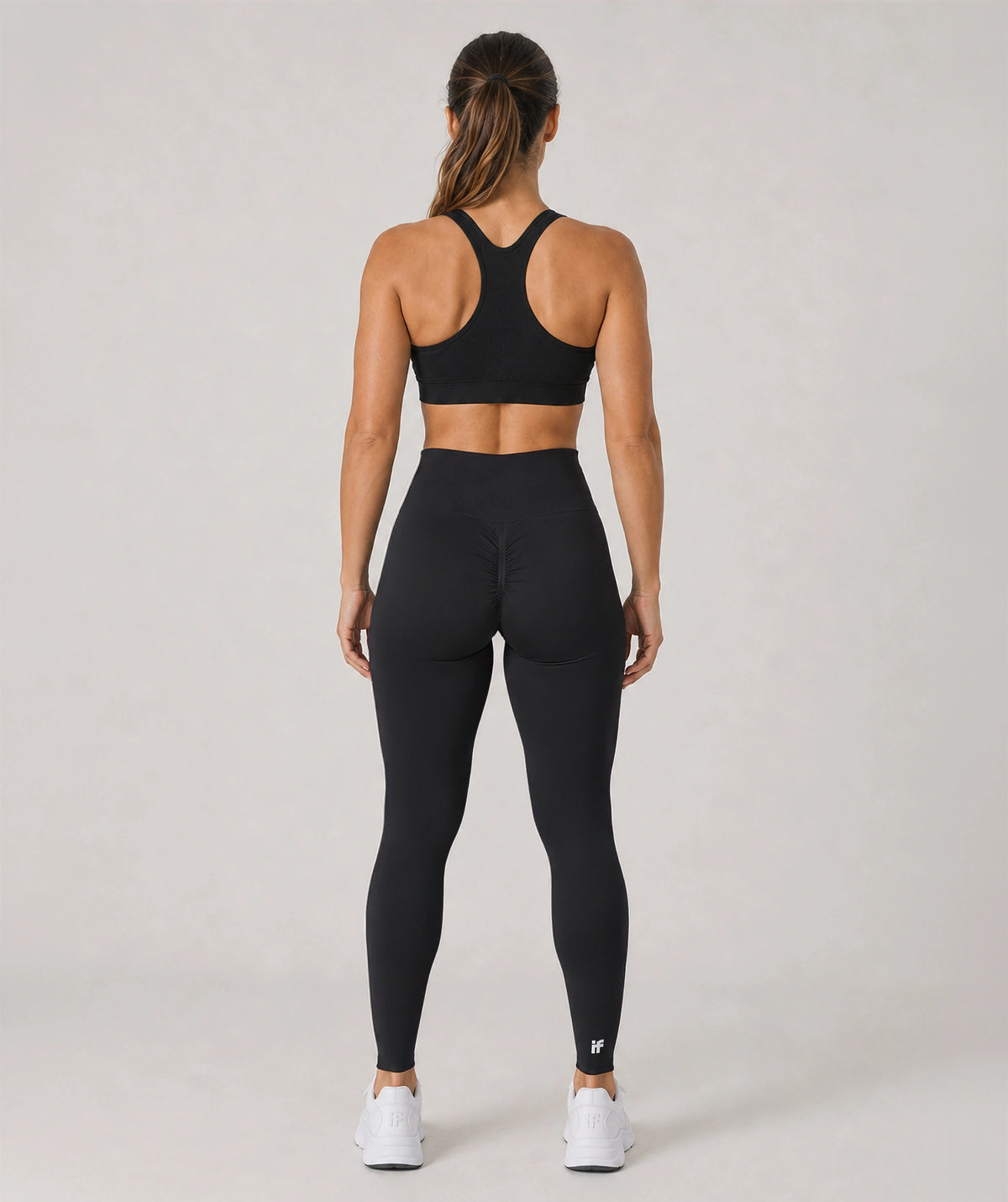 A woman viewed from the back wearing a black sports bra and black high-waisted leggings, paired with white sneakers, against a light gray studio background.