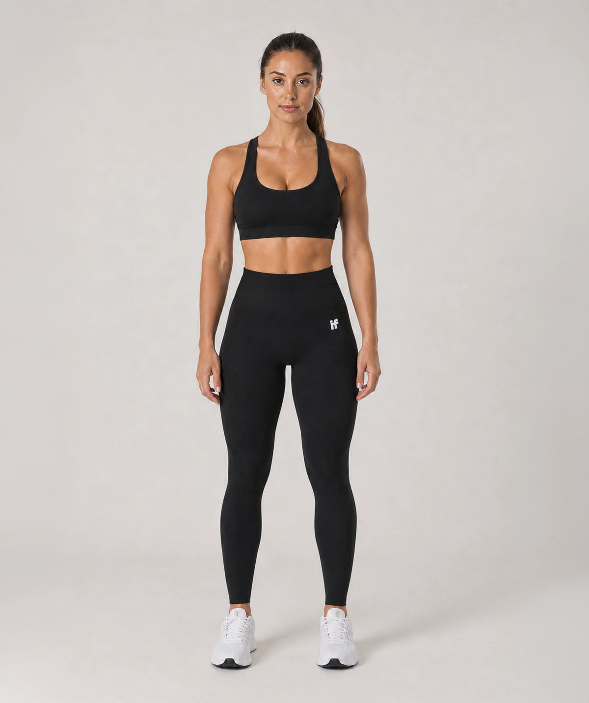 A woman modeling a black athletic sports bra and matching high-waisted leggings with a small white logo on the left hip, paired with white sneakers, against a light gray studio background.