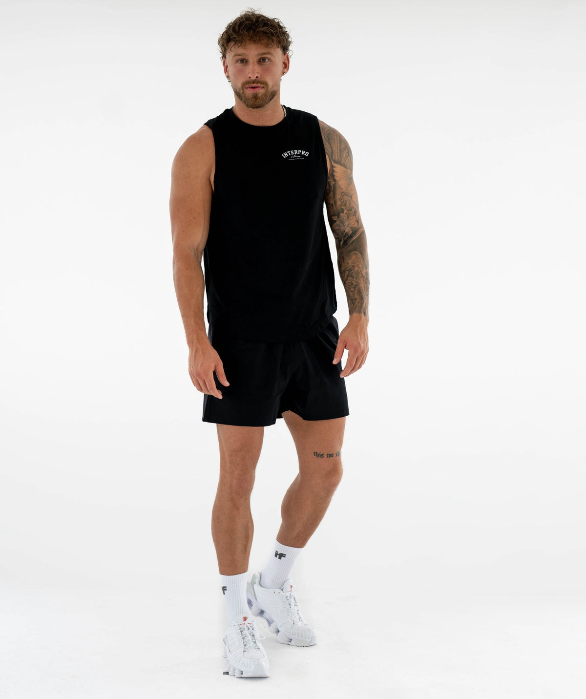 A man modeling a black sleeveless athletic tank top and matching black shorts against a plain white studio background. The tank top has a small white logo on the left chest, and the model is also wearing white socks and white sneakers.