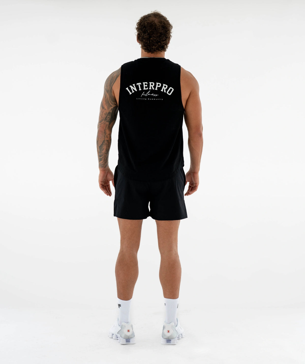 A man viewed from behind wearing a black sleeveless athletic top and matching black shorts, with white socks and white sneakers, against a clean white studio background. The top has 'INTERPRO' printed in white across the upper back.